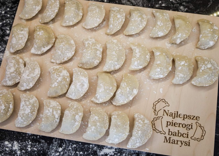 Drewniana deska z grawerem pierogów i napisem: "Najlepsze pierogi babci Marysi", na desce leżą pierogi