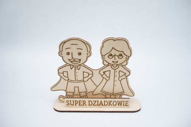 Drewniana statuetka z postaciami babci i dziadka jako super bohaterowie, przed nimi napis: super dziadkowie, figurka i napis na podstawce