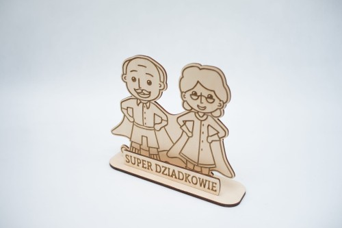 Drewniana statuetka z postaciami babci i dziadka jako super bohaterowie, przed nimi napis: super dziadkowie, figurka i napis na podstawce